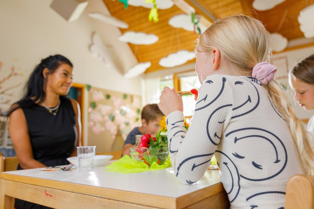 Eine Erzieherin sitzt am Tisch mit essenden Kindern.